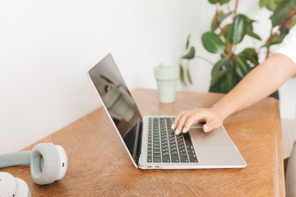 A hand using a laptop on a wooden desk with headphones and a green plant nearby, depicting modern remote work.
